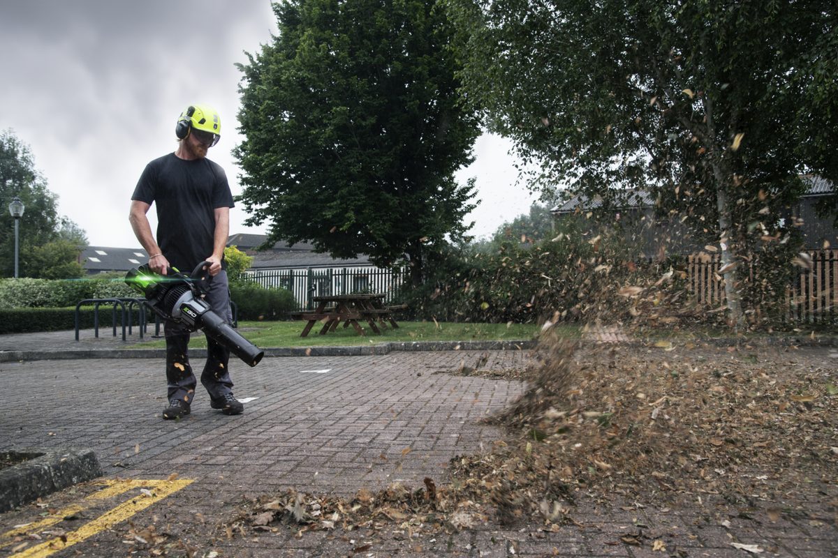 Kutseline töötaja kiivri, kuulmiskaitse ja kaitseprillidega puhub EGO LBX1000 lehepuhuriga lehti parklast