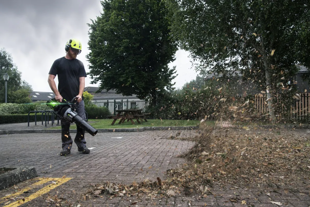Kutseline töötaja kiivri, kuulmiskaitse ja kaitseprillidega puhub EGO LBX1000 lehepuhuriga lehti parklast
